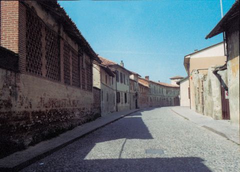 Via Corridoni fiancheggiata dalle cascine; in primo piano la strada acciottolata; sullo sfondo, il castello Brivio.